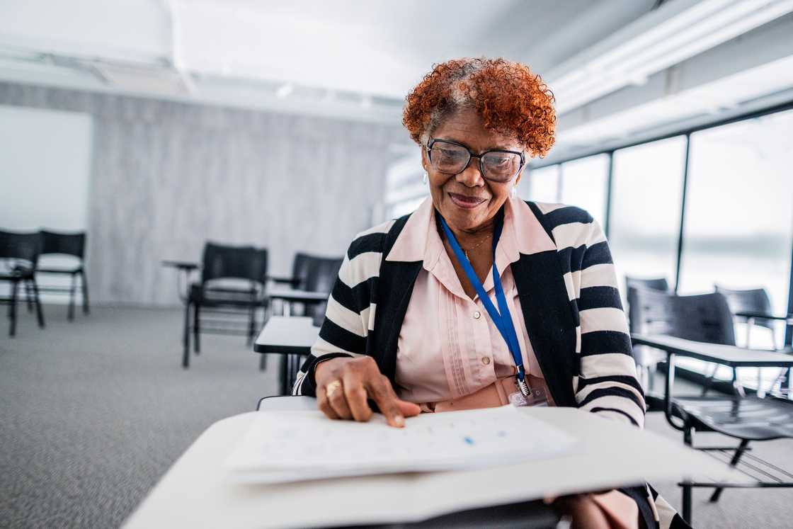 Older adult in glasses points at a worksheet while seated in a bright classroom.