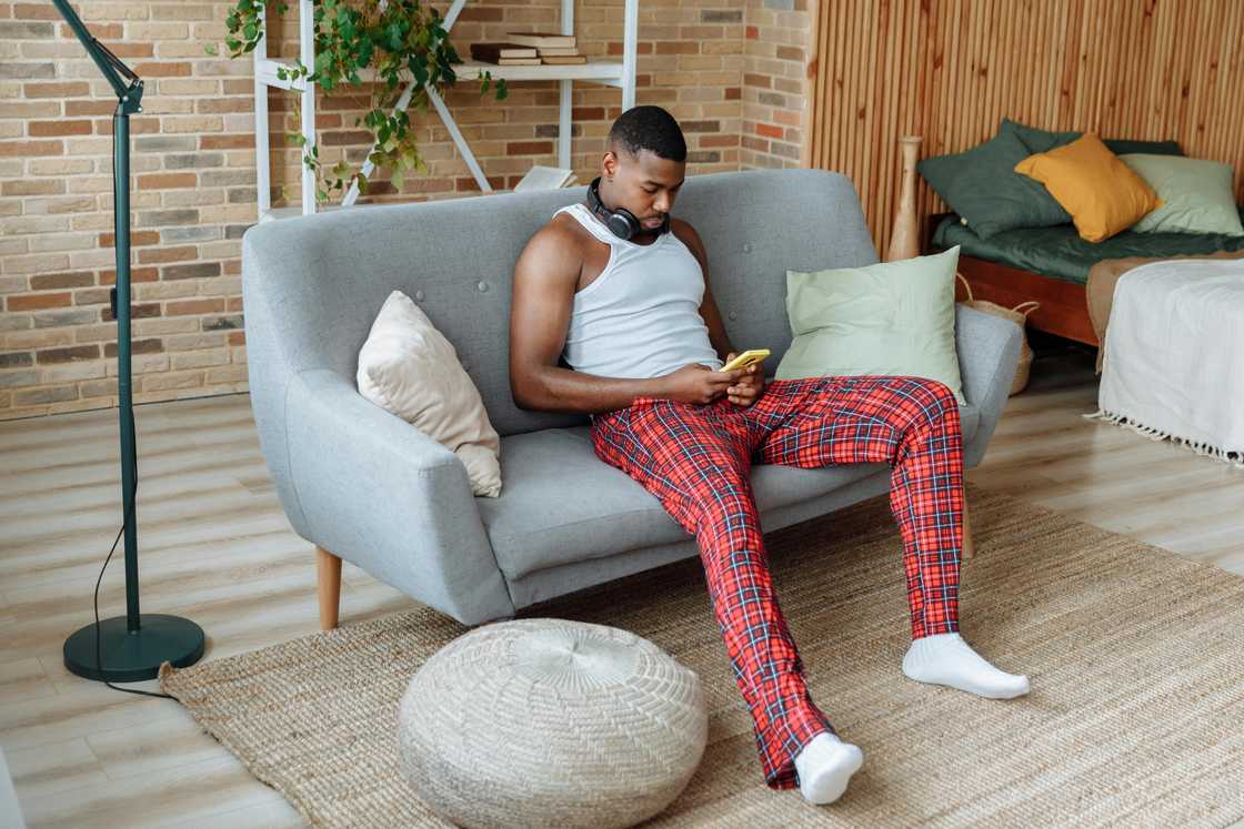 A man in a white vest and checkered trousers is using a smartphone on a grey sofa