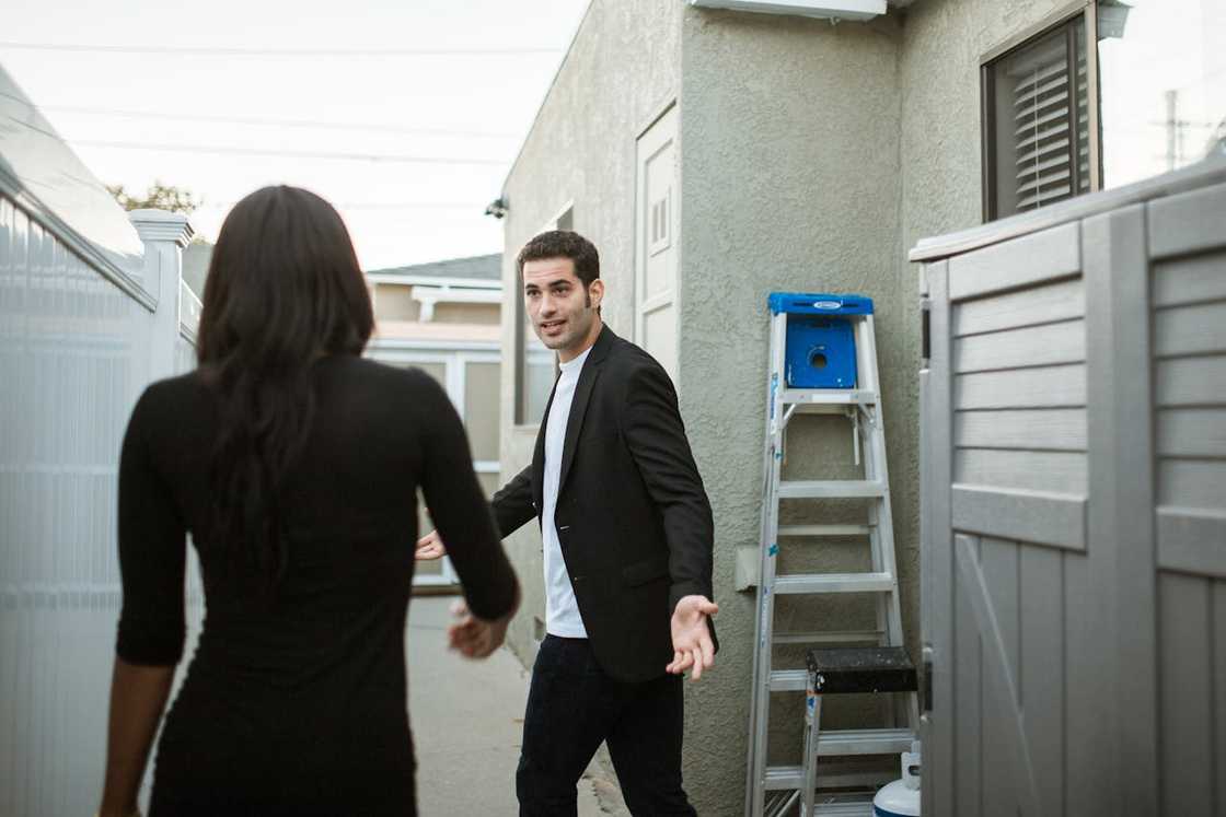 A man gestures toward a woman during a tense exchange in a narrow outdoor walkway.
