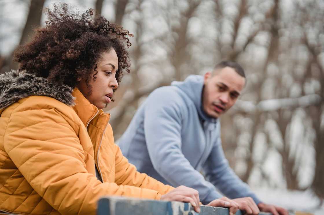 A woman sits pensively on a bench while a man looks at her outdoors.