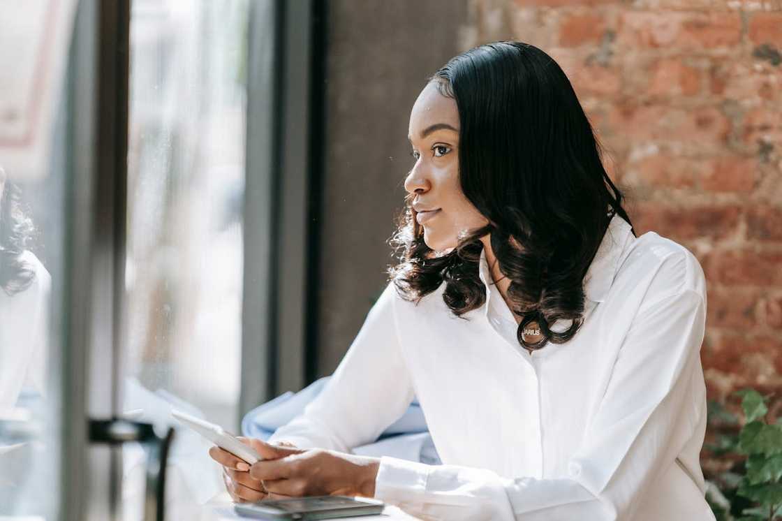 A woman sits by a window holding a phone and looking outside thoughtfully. A woman sits by a window holding a phone and looking outside thoughtfully.