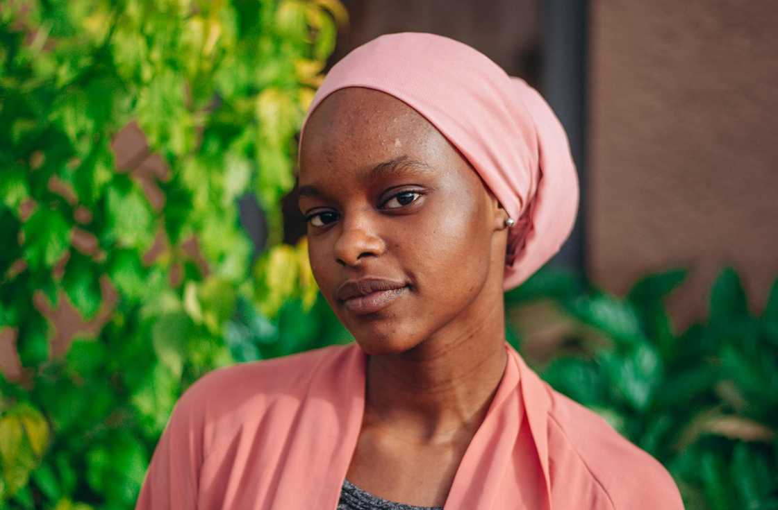 A woman wearing a pink headscarf looks calmly at the camera outdoors.