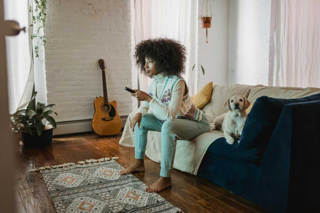 A woman in blue pants is watching the TV next to her dog on a navy blue couch with a brown throw blanket