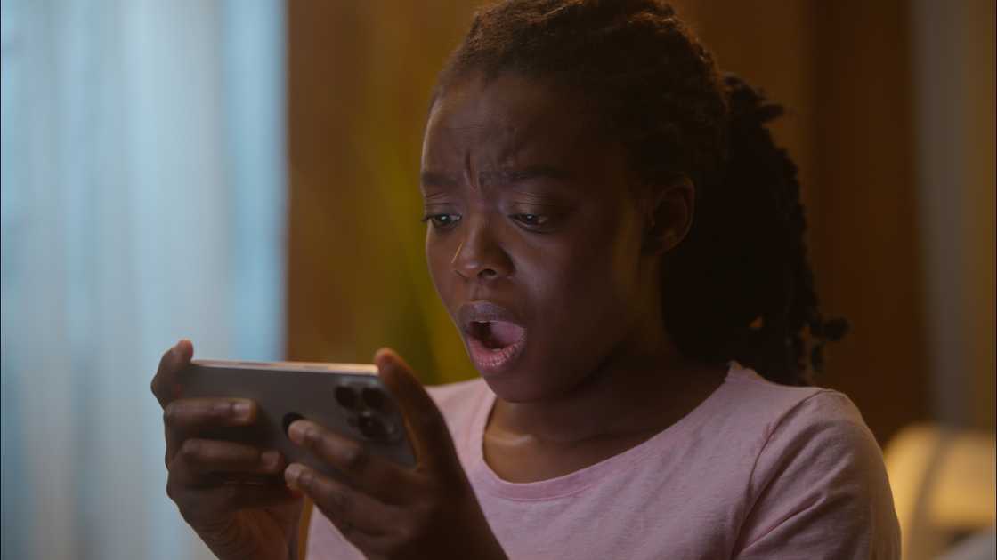 A person in a light pink shirt reacts with wide eyes and open mouth while looking at a smartphone. A person in a light pink shirt reacts with wide eyes and open mouth while looking at a smartphone.