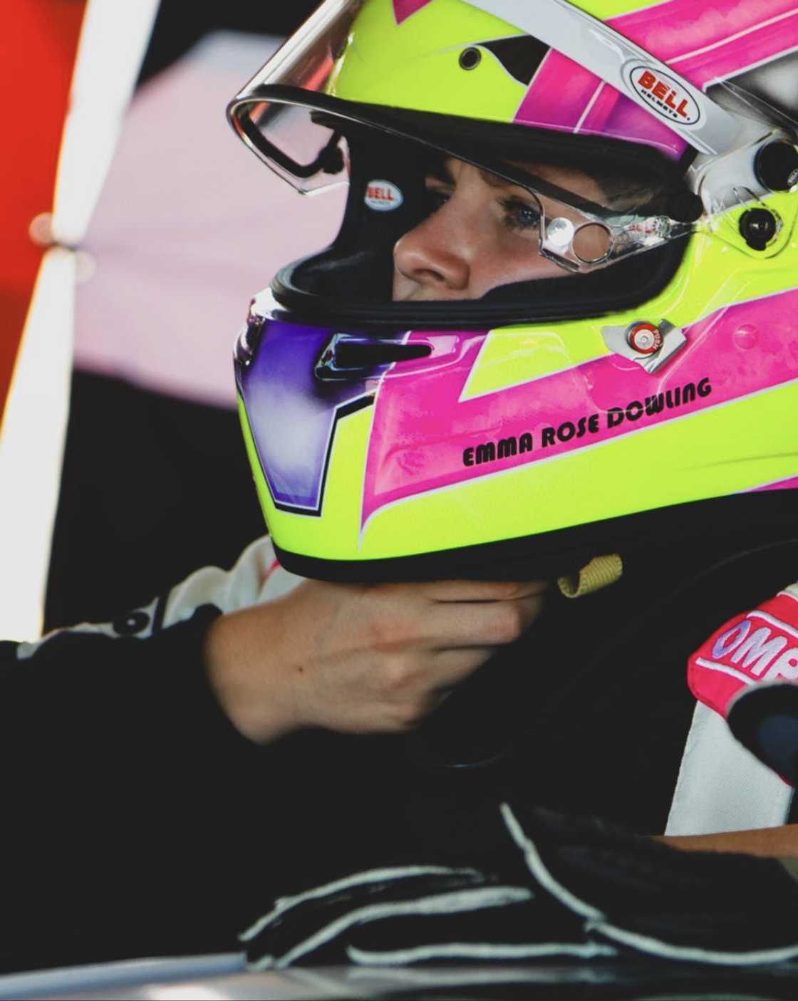 Close-up of Emma-Rose Dowling adjusting her pink and neon yellow Bell helmet, with her name printed on the side, while seated in her race car and wearing racing gloves.