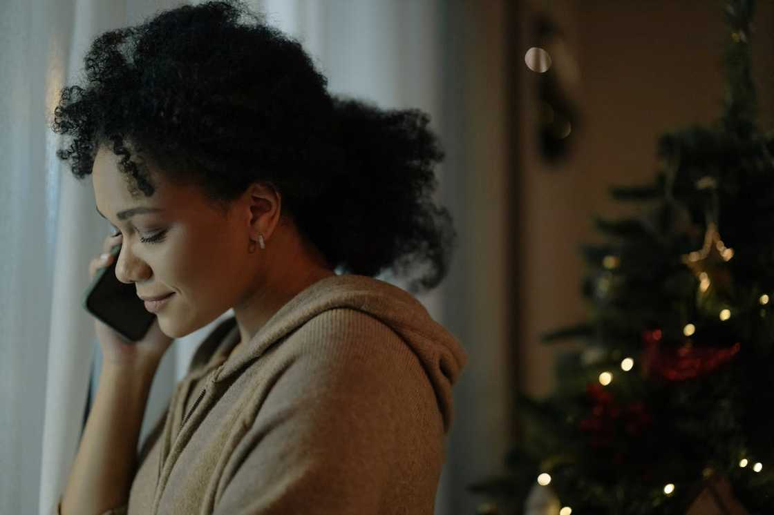 A woman stands near a window smiling softly, while talking on her phone beside a lit Christmas tree.