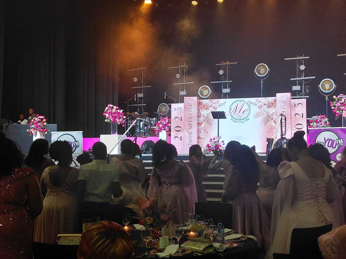 Women in elegant pink dresses gather at the Me Time Event, with a stage backdrop showcasing the 2025 theme "I Am Every Woman" in a lively and inspiring atmosphere