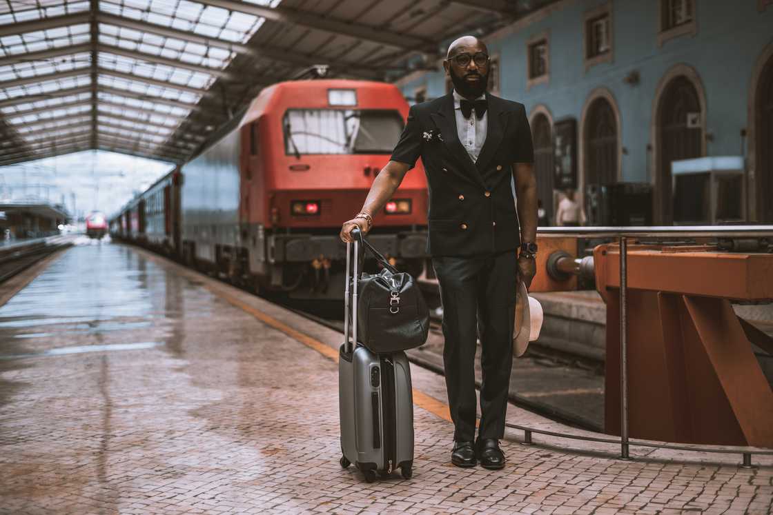 Person in formal attire stands on a train platform beside a silver suitcase and black bag. Person in formal attire stands on a train platform beside a silver suitcase and black bag.