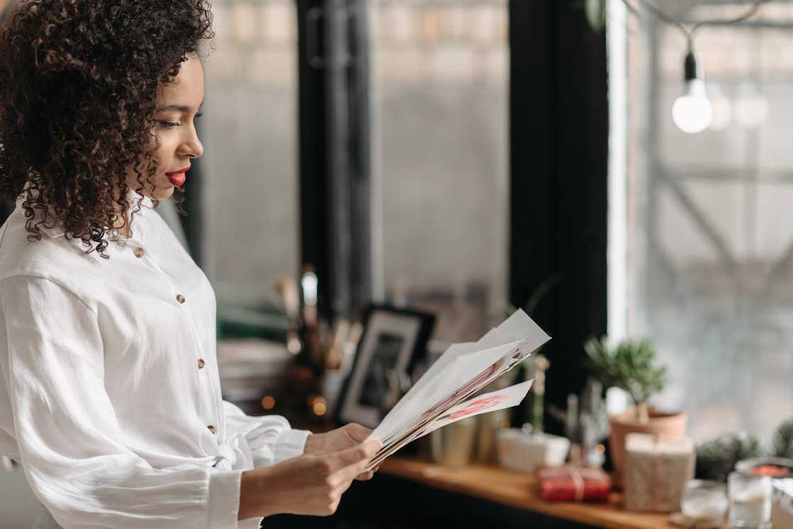 A woman looks at several papers in her hands while standing near a window.