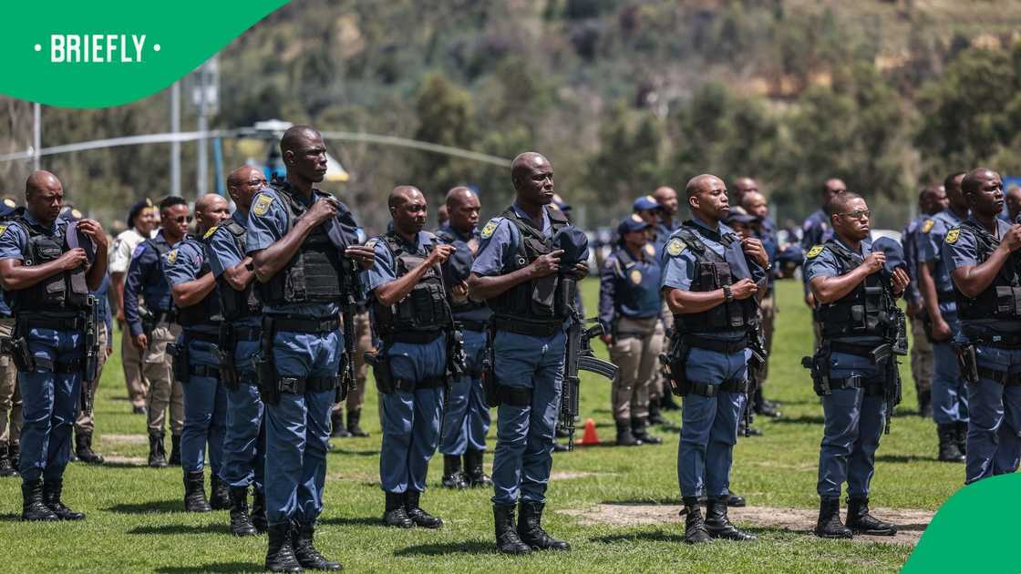 A line of South African Police Service officers standing at rigid attention in their dark blue field dress uniforms, wearing matching caps and duty belts during an outdoor ceremonial parade.