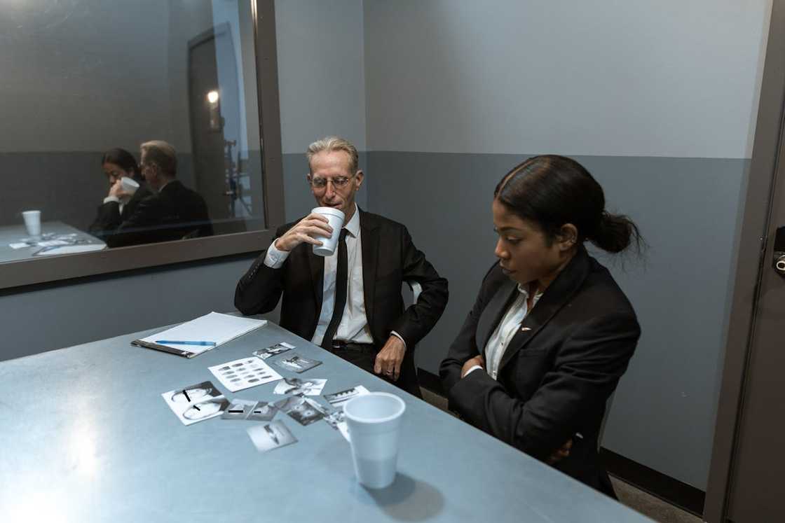 A woman and a man seated at a table in a stark interview room. A woman and a man seated at a table in a stark interview room.