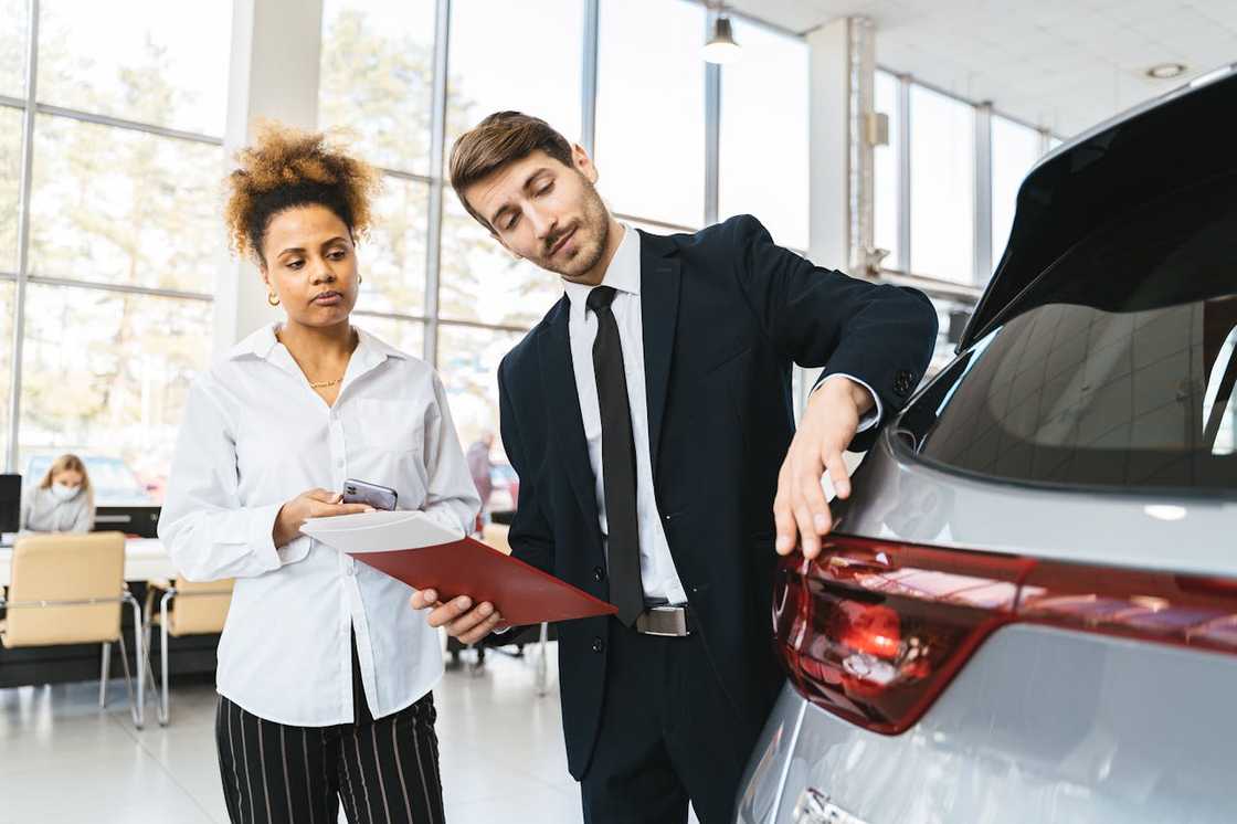 A car inspector showing a car to a woman while reviewing documents.