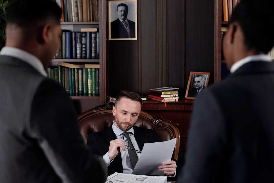 A lawyer reading paperwork in an office with two people standing nearby. A lawyer reading paperwork in an office with two people standing nearby.