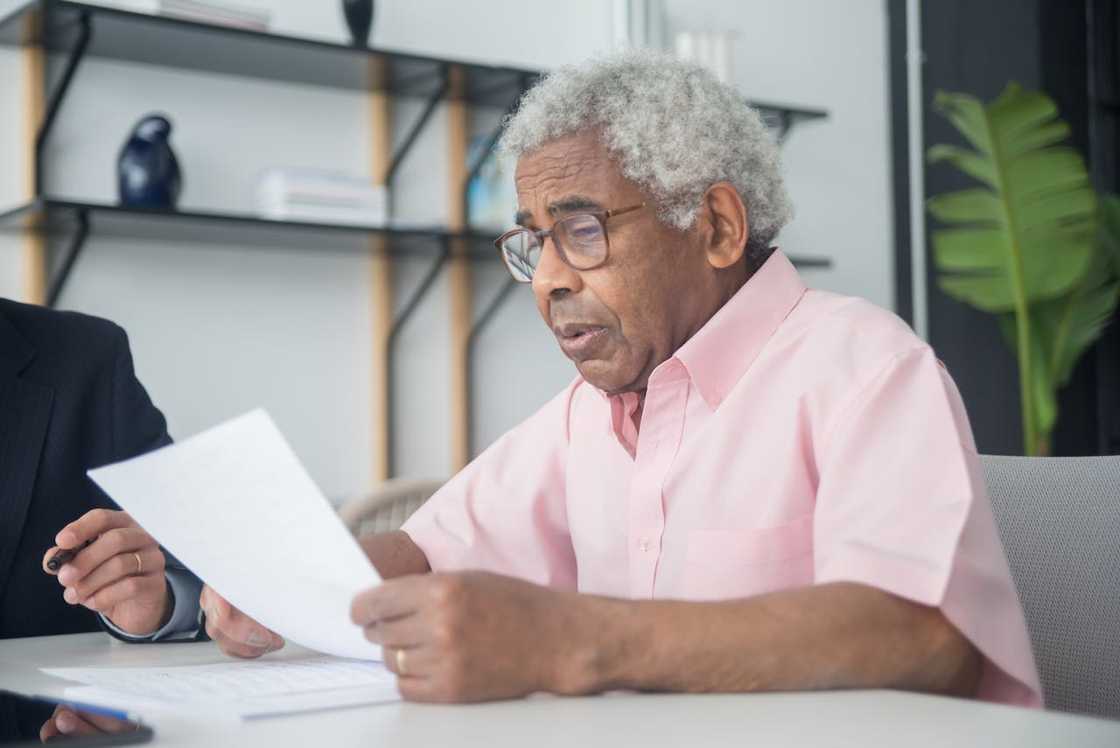 An elderly man reads a document while seated at a desk in an office. An elderly man reads a document while seated at a desk in an office.