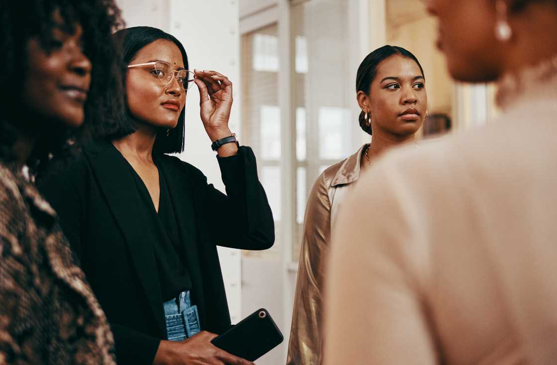 Two people stand indoors, one adjusting glasses while holding a phone. Two people stand indoors, one adjusting glasses while holding a phone.