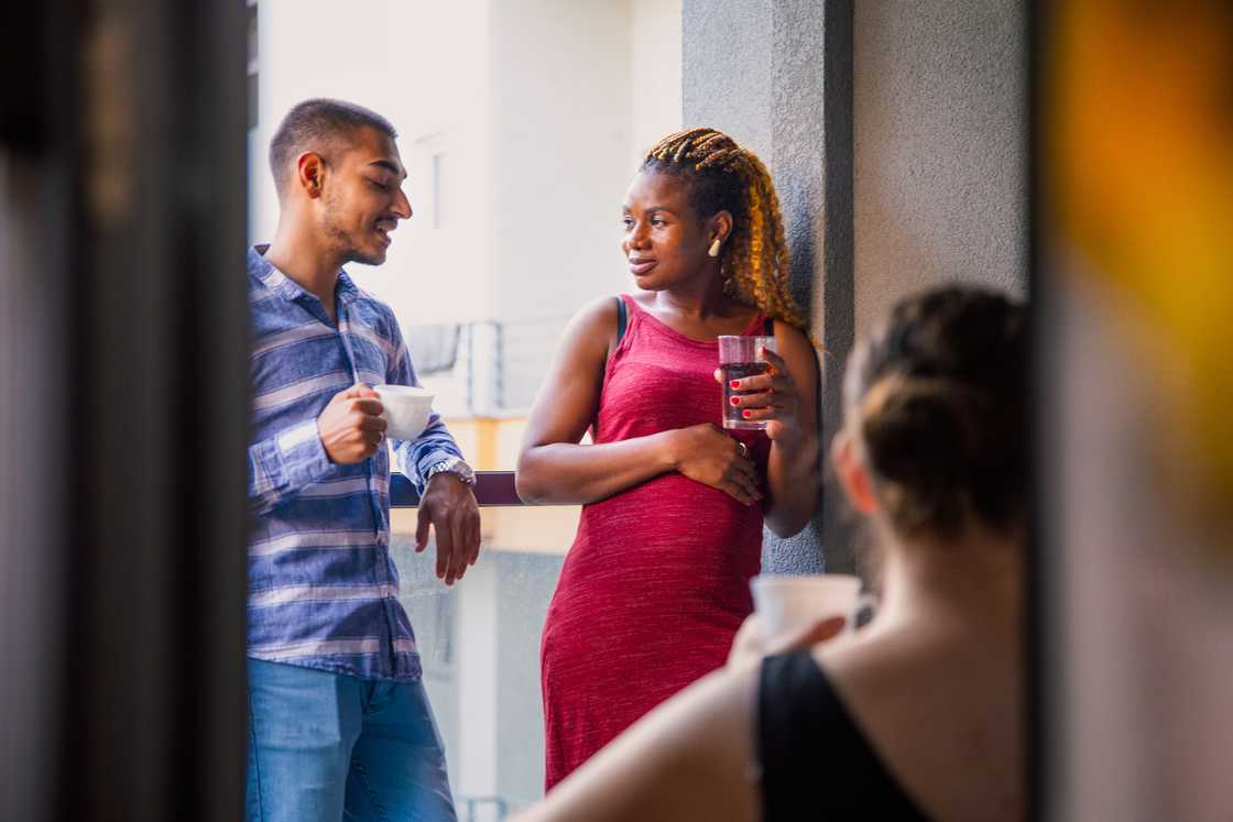 Three people on a balcony, two talking, one seated with a cup. Three people on a balcony, two talking, one seated with a cup.