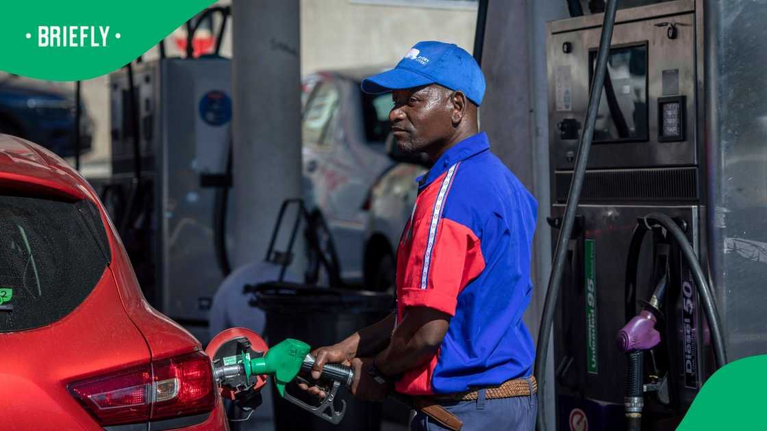A fuel station attendant wearing a blue cap and a blue and red Engen uniform inserts a green petrol pump nozzle into the fuel tank of a red car at a service station forecourt.