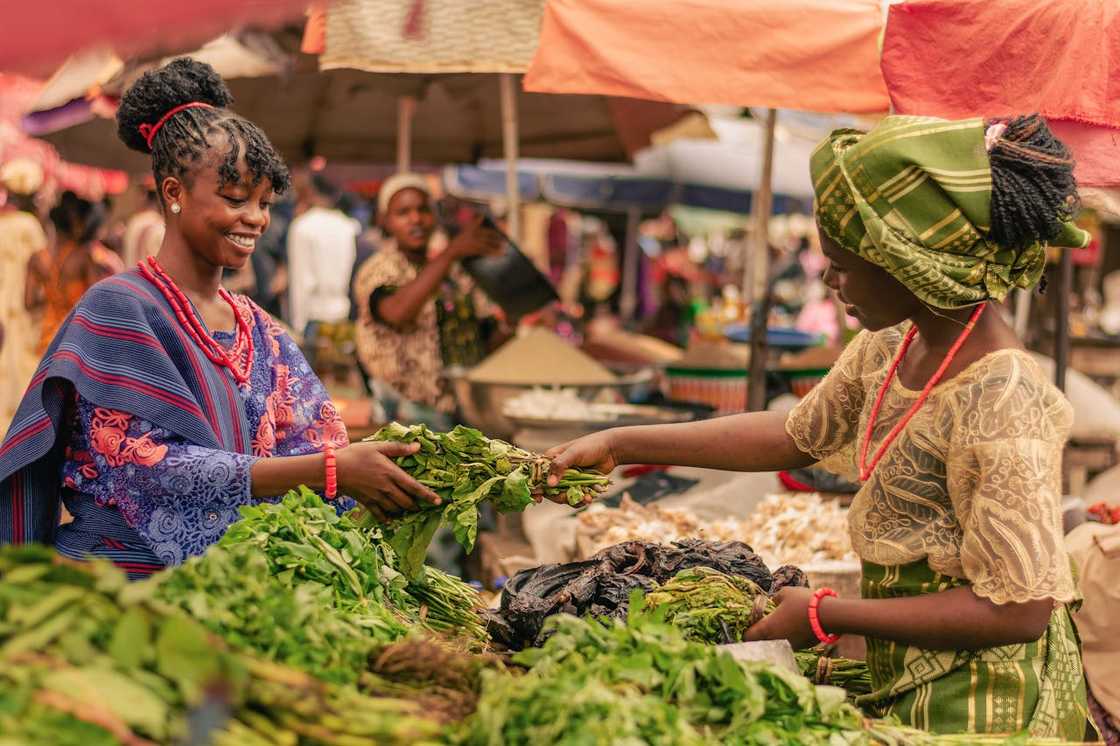 Two women exchange leafy vegetables at an outdoor market.