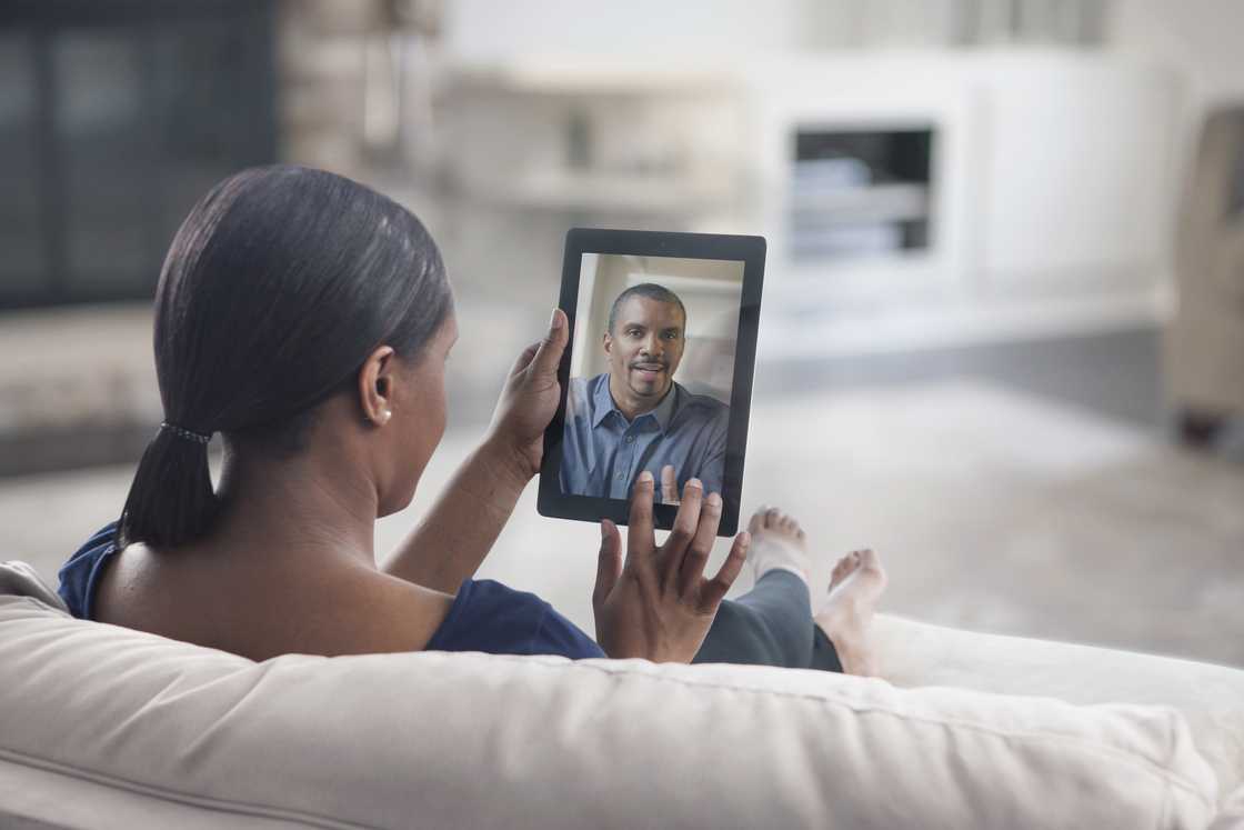 Person sits on a couch holding a tablet, engaged in a video call with someone onscreen.