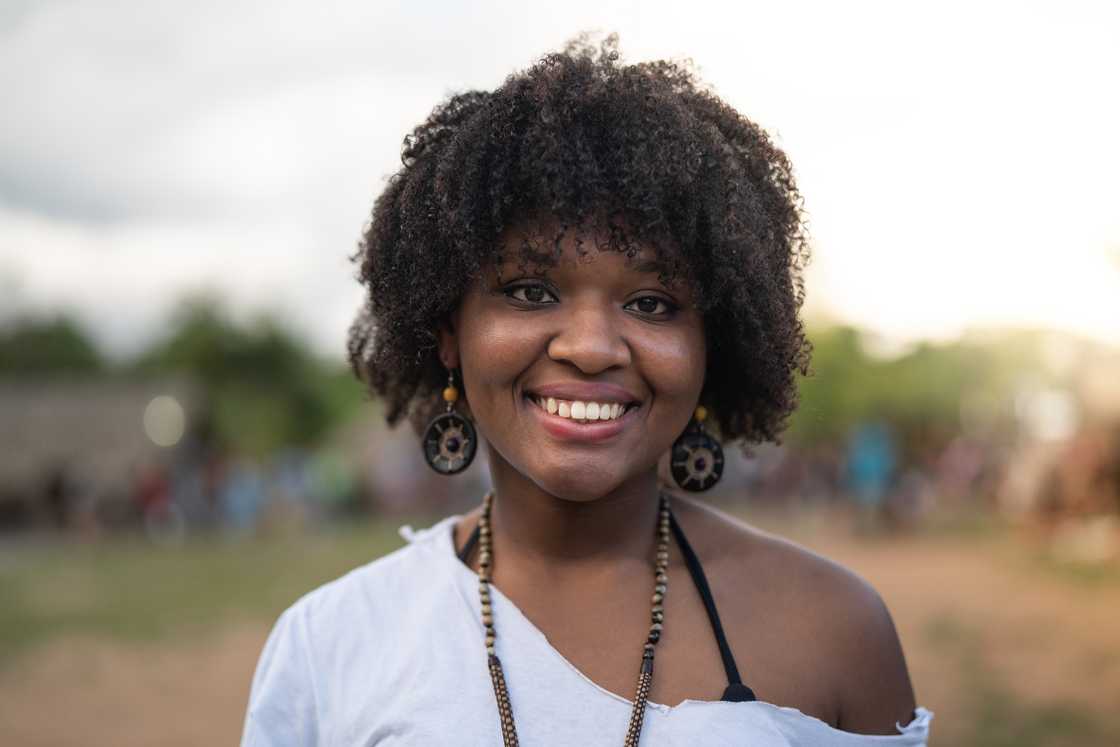 Smiling woman in a white off-shoulder top stands outdoors with greenery in the background.