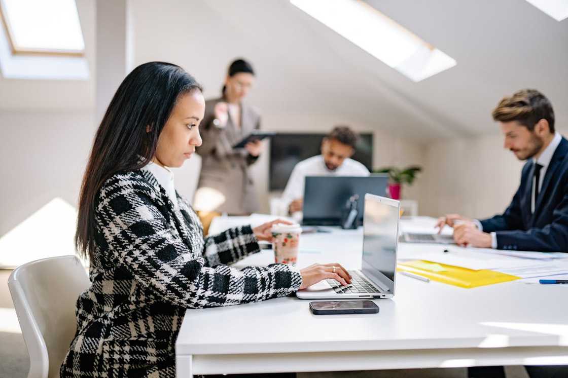 A woman working on a laptop at a shared office table while colleagues appear in the background.