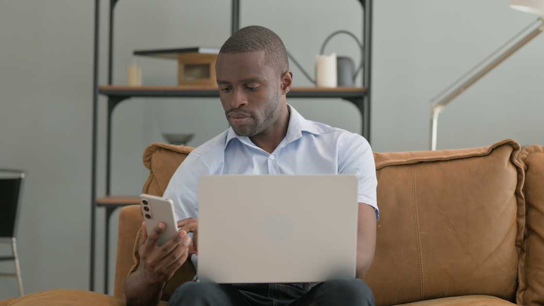 A man using a laptop and smartphone while sitting on a sofa at home A man using a laptop and smartphone while sitting on a sofa at home