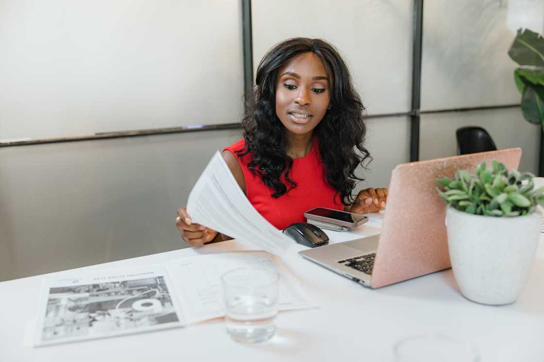 A woman in a red top works at a desk with a laptop and documents.