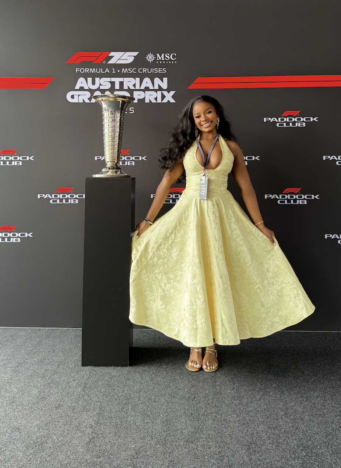 Pamela Mtanga smiles beside the Austrian Grand Prix trophy, wearing a yellow gown and F1 Paddock Club lanyard.