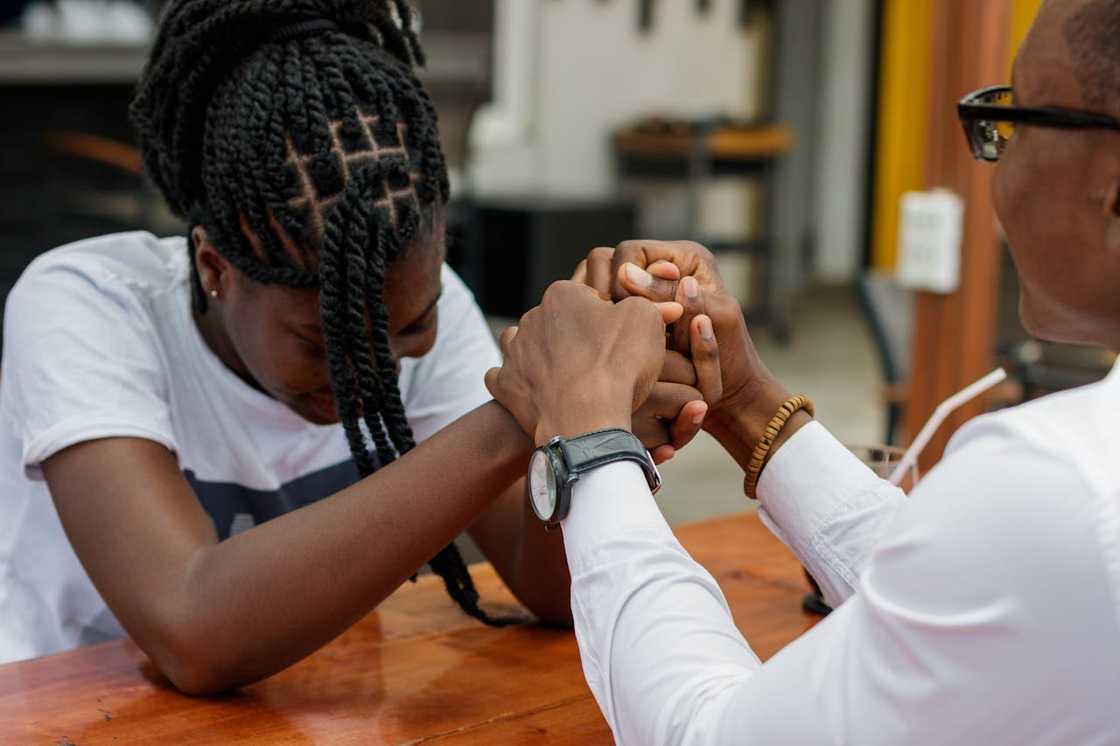 Two people sit at a table holding hands in a moment of emotional support. Two people sit at a table holding hands in a moment of emotional support.