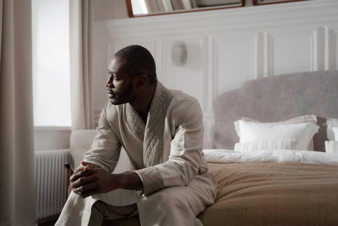 A man sitting on a bed, looking out a window in a quiet bedroom.