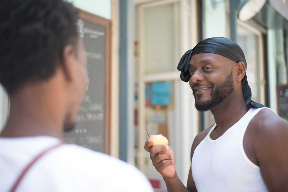 A man smiles while holding a pear and talking to another person outdoors.