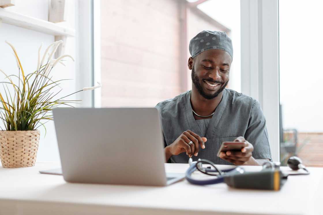 A happy man in grey scrubs is using his smartphone next to a table with a silver laptop