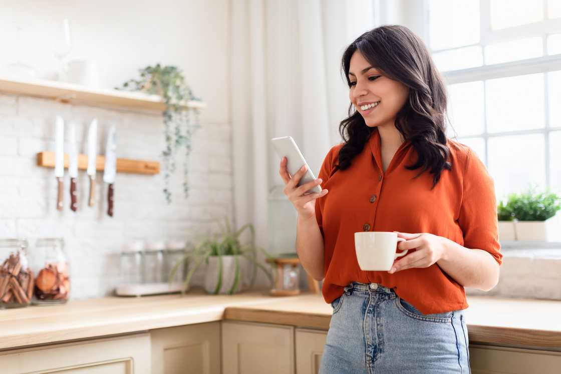 A woman in her kitchen smiling at her phone.