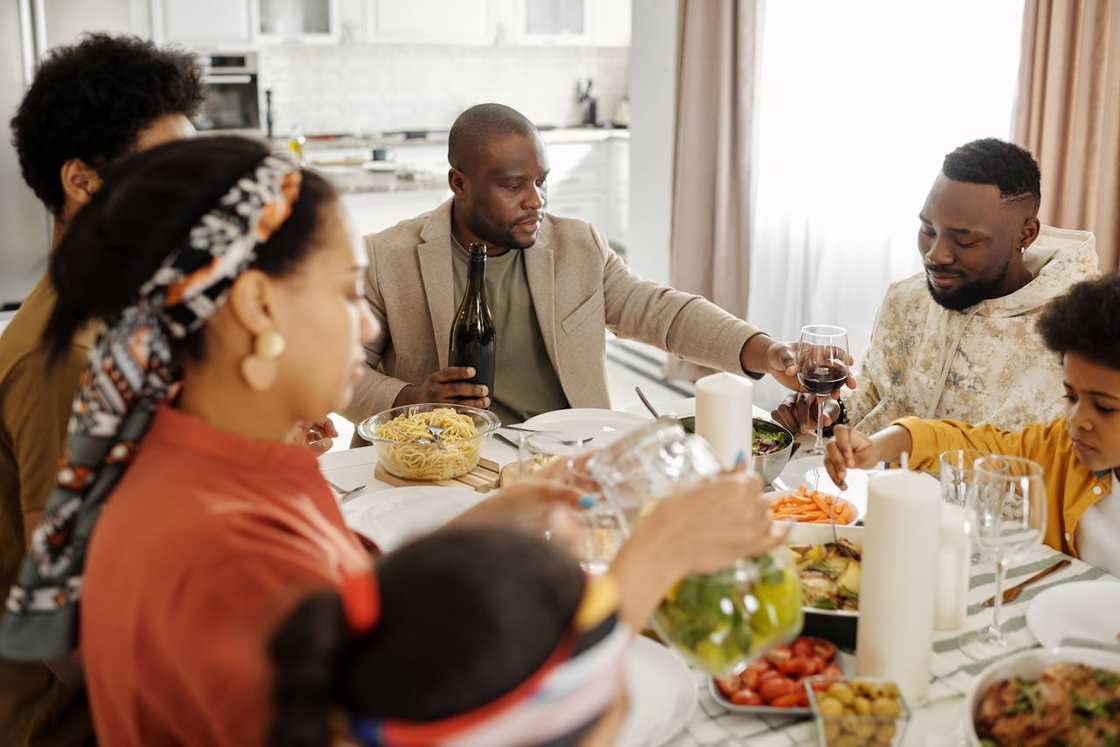 A family eats together at a dining table while a man pours wine for another guest.