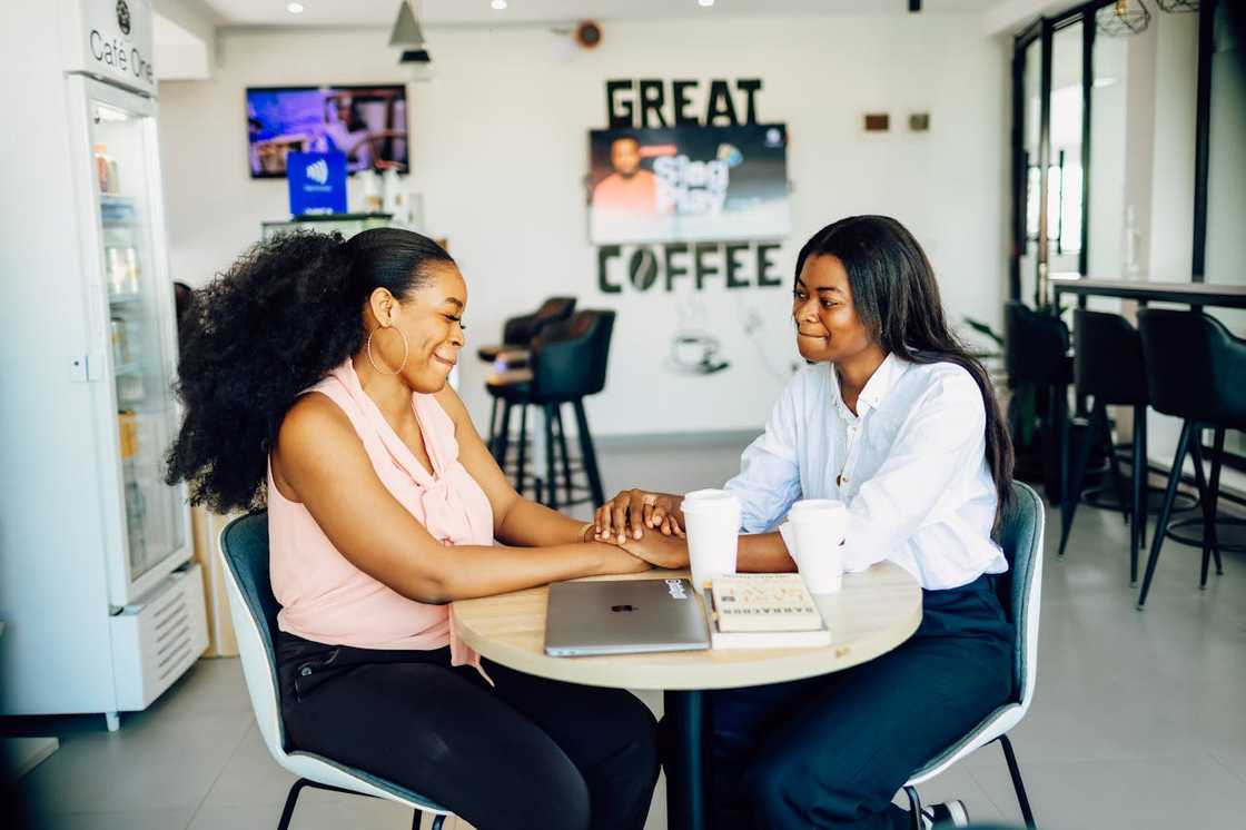 Two women sit at a café table, holding hands and talking over coffee.