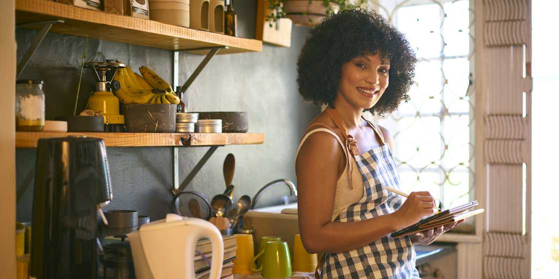 Person in a checkered apron smiles while writing in a notebook in a cozy kitchen. Person in a checkered apron smiles while writing in a notebook in a cozy kitchen.