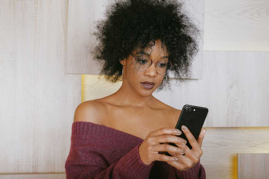 A woman wearing glasses looks down at her phone while standing against a light wooden wall.