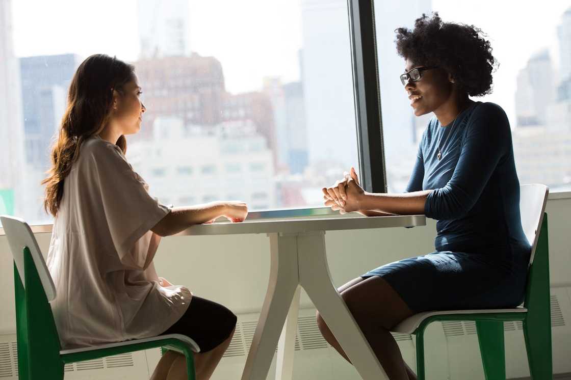 Two women sitting across a table by a window, engaged in a serious conversation.