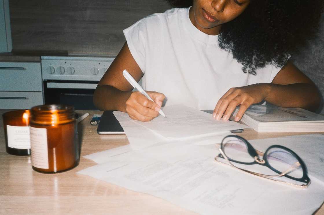 A woman writing in a notebook at a table with papers, glasses, and lit candles nearby.