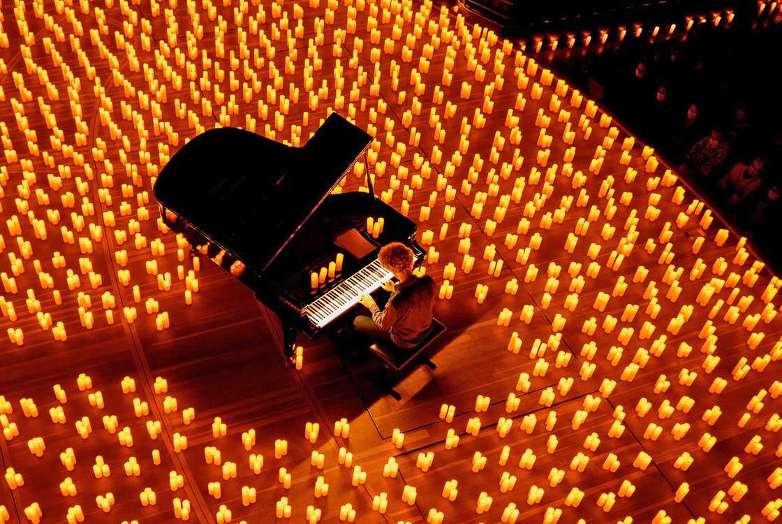 An overhead view of a pianist playing a black grand piano on a wooden stage completely surrounded by thousands of glowing, warm amber candles.