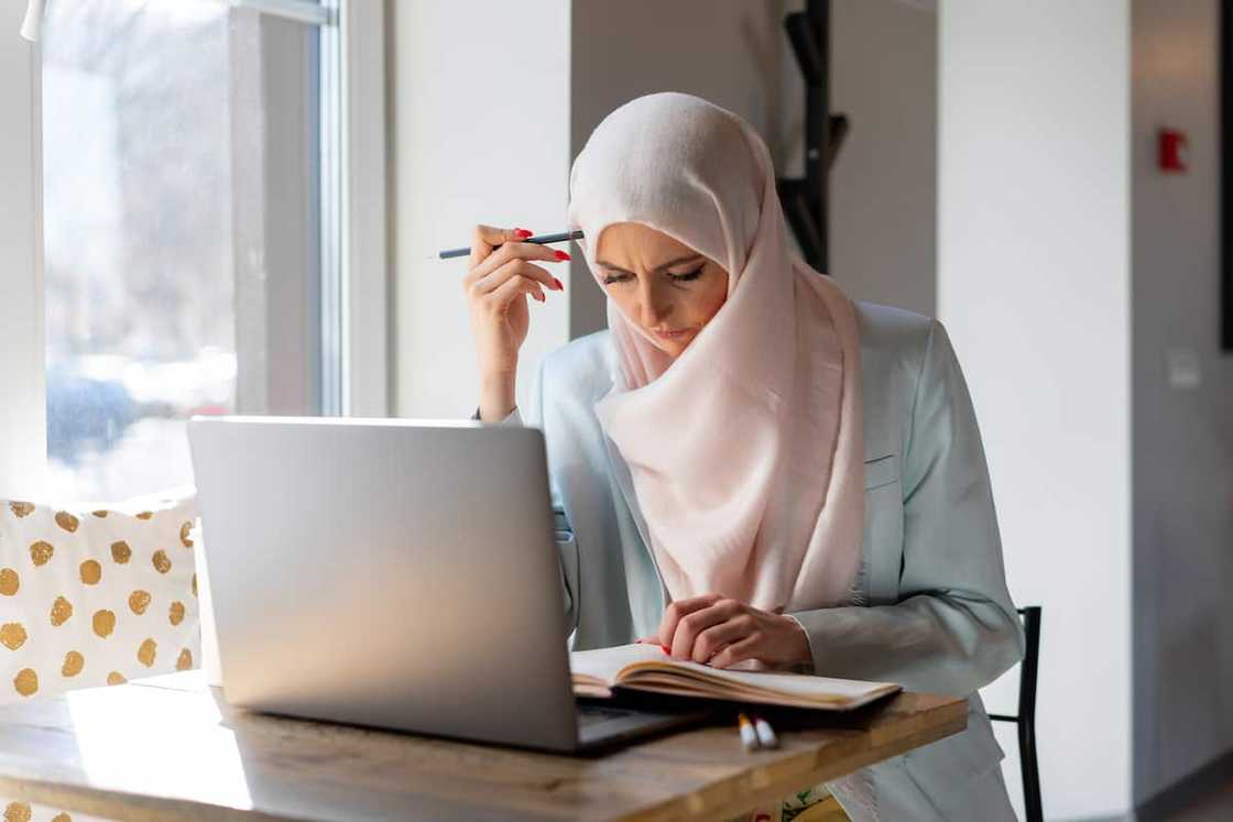 A woman reading a book with a laptop on her desk