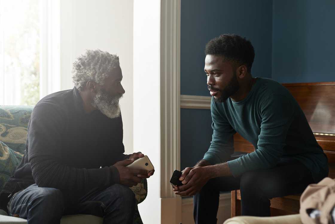 An older man converses with a younger man in a home setting. An older man converses with a younger man in a home setting.