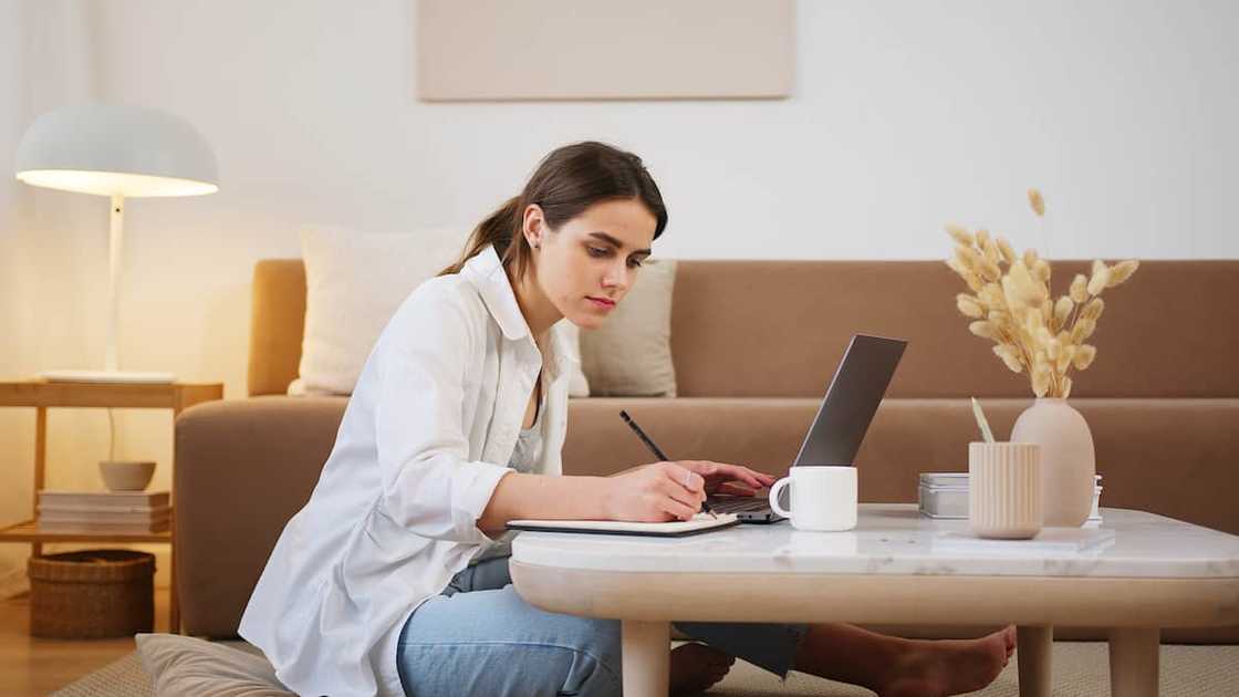 A lady sitting on the floor of her living while taking notes from a laptop.