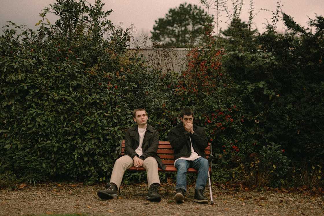 Two men sit on a bench outdoors, looking tense and quiet.