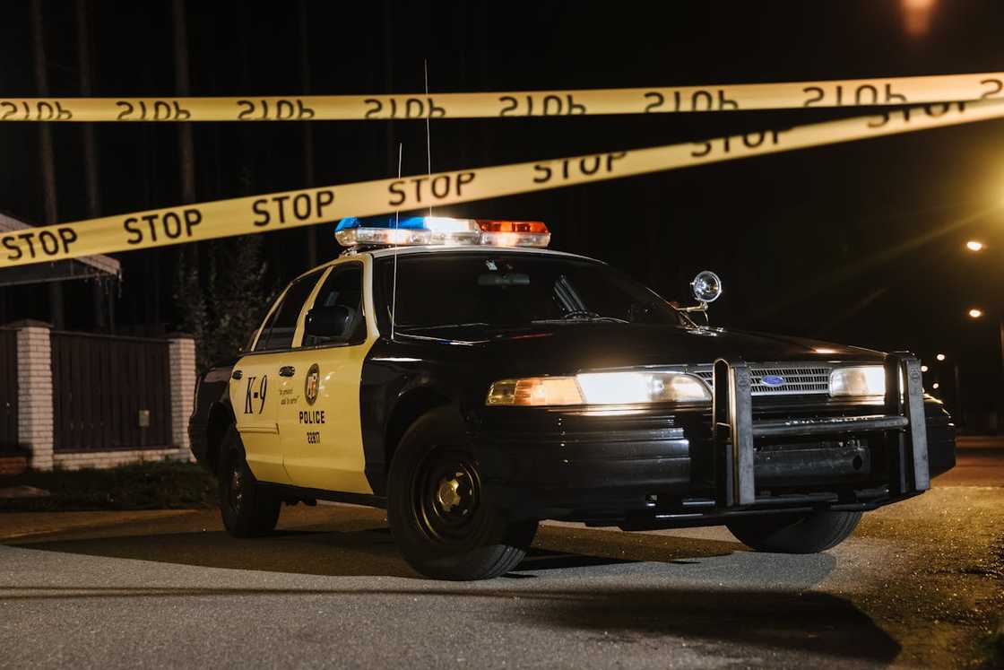 A police car parked at night behind yellow “STOP” crime-scene tape. A police car parked at night behind yellow “STOP” crime-scene tape.