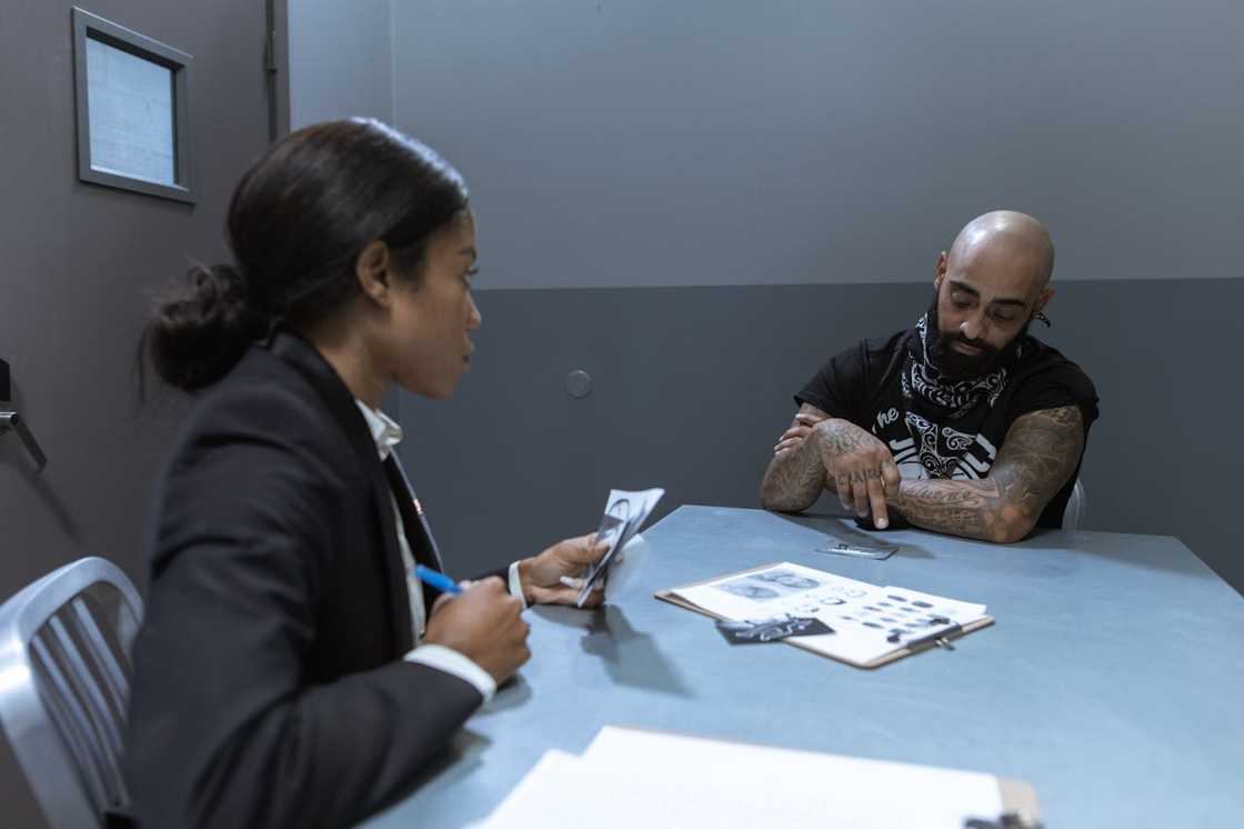 An investigator questions a tattooed man across a metal table. An investigator questions a tattooed man across a metal table.