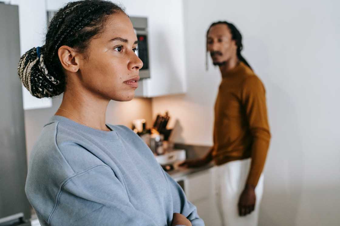 A woman stands apart while her partner watches from the kitchen.
