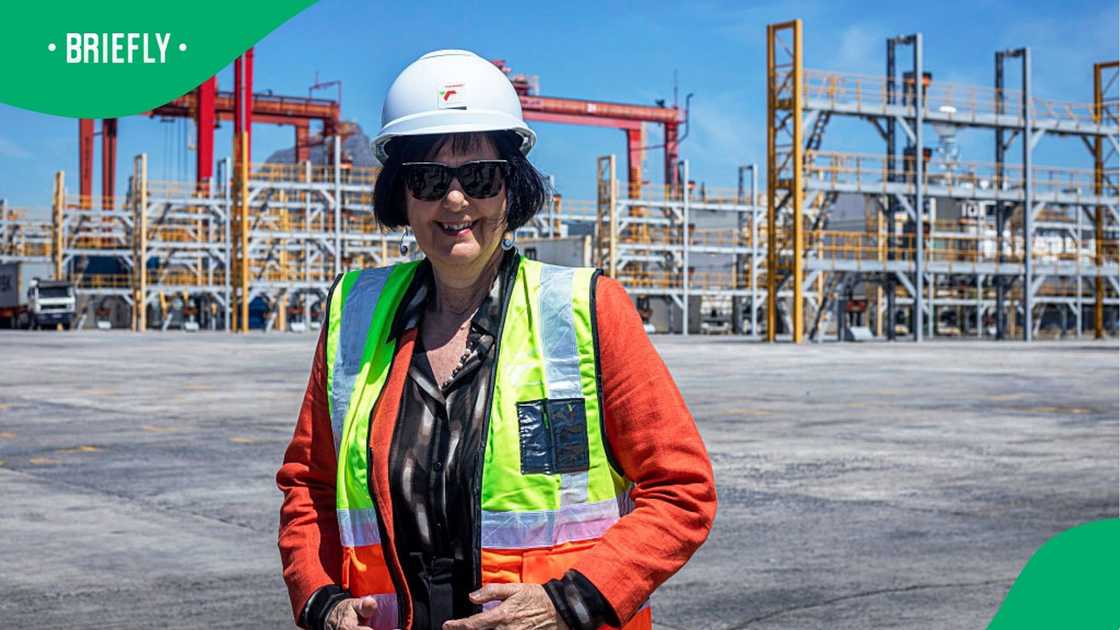 The minister stood outdoors on a concrete tarmac with large shipping cranes and harbour infrastructure visible in the background