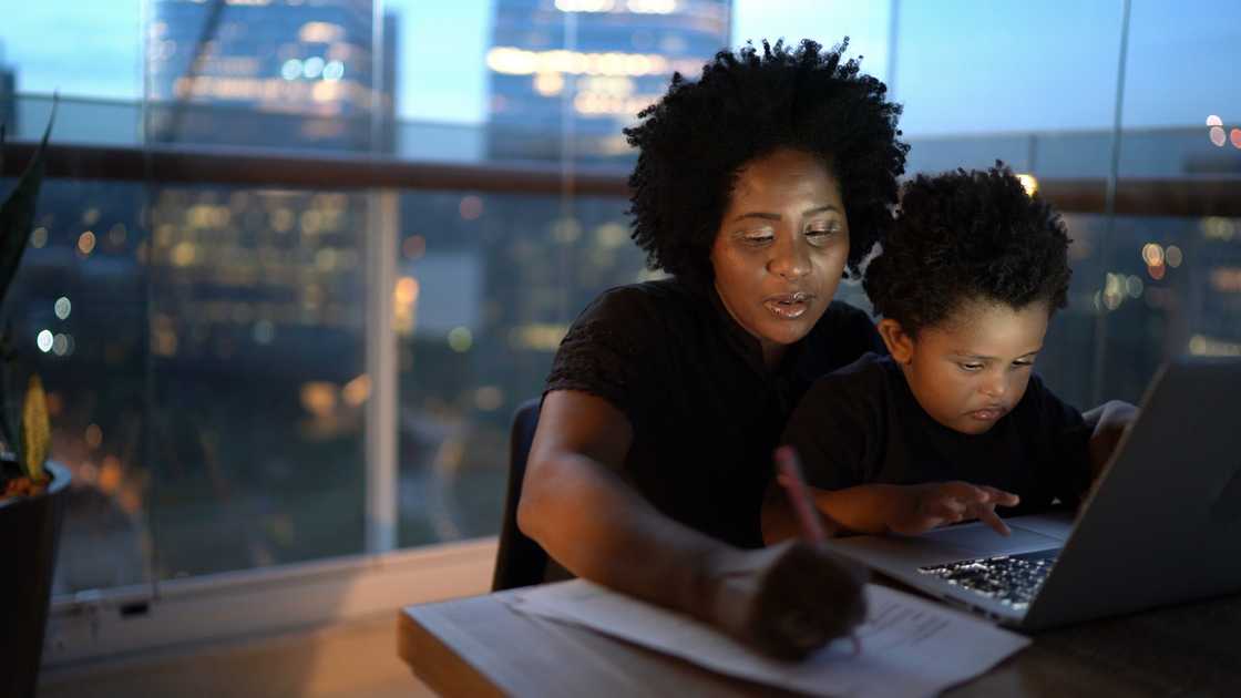 Adult and child sit at a table with a laptop, working together as city lights glow outside.