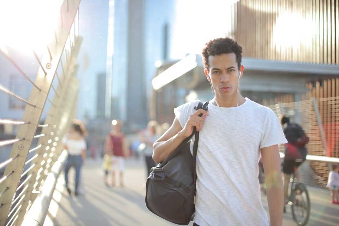 A young man walks outdoors with a backpack over his shoulder, listening to earphones.